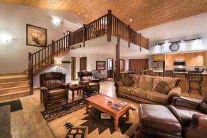 A spacious living area with a high wooden ceiling, a staircase, leather furniture, and a kitchen in the background.