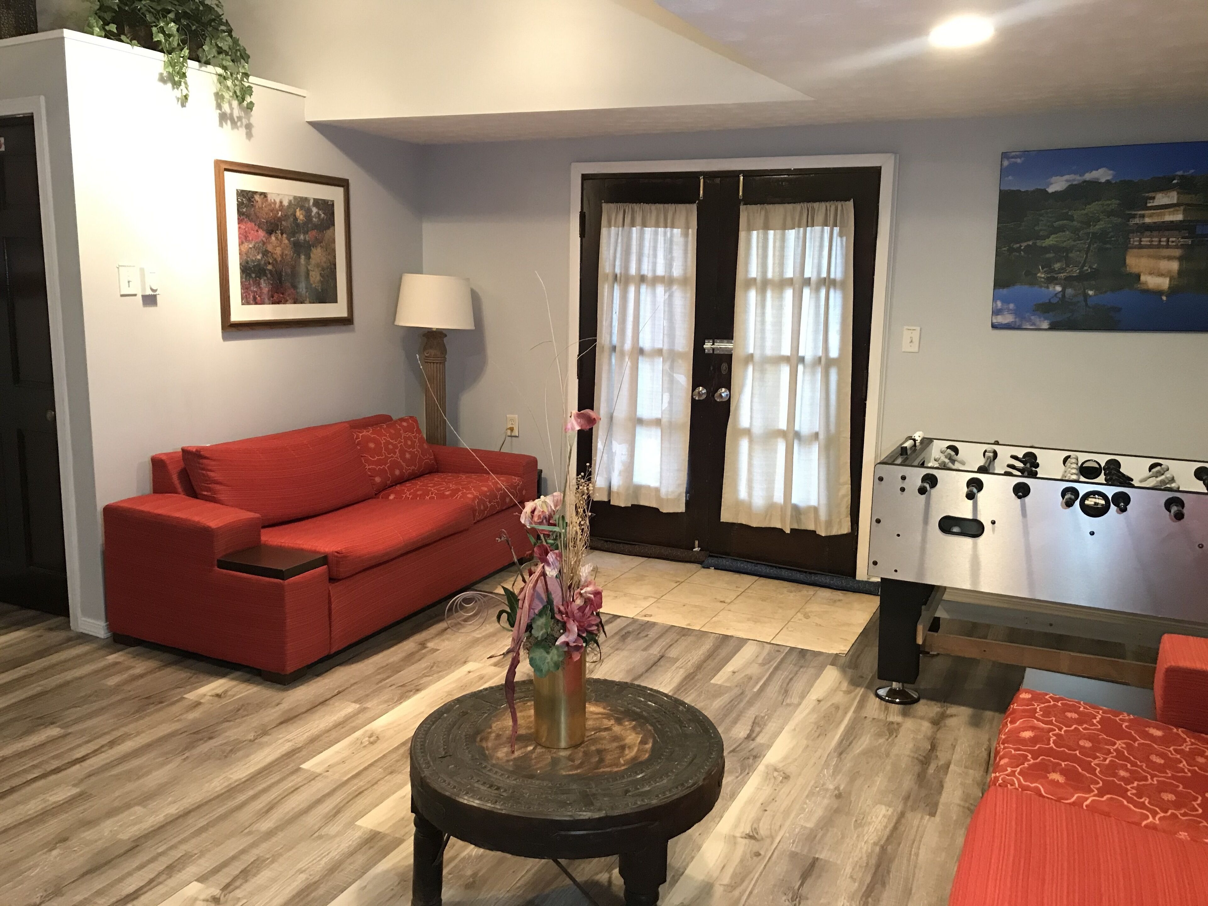 A living room with a red sofa, a round coffee table with a vase of flowers, a foosball table, and a door with curtains.
