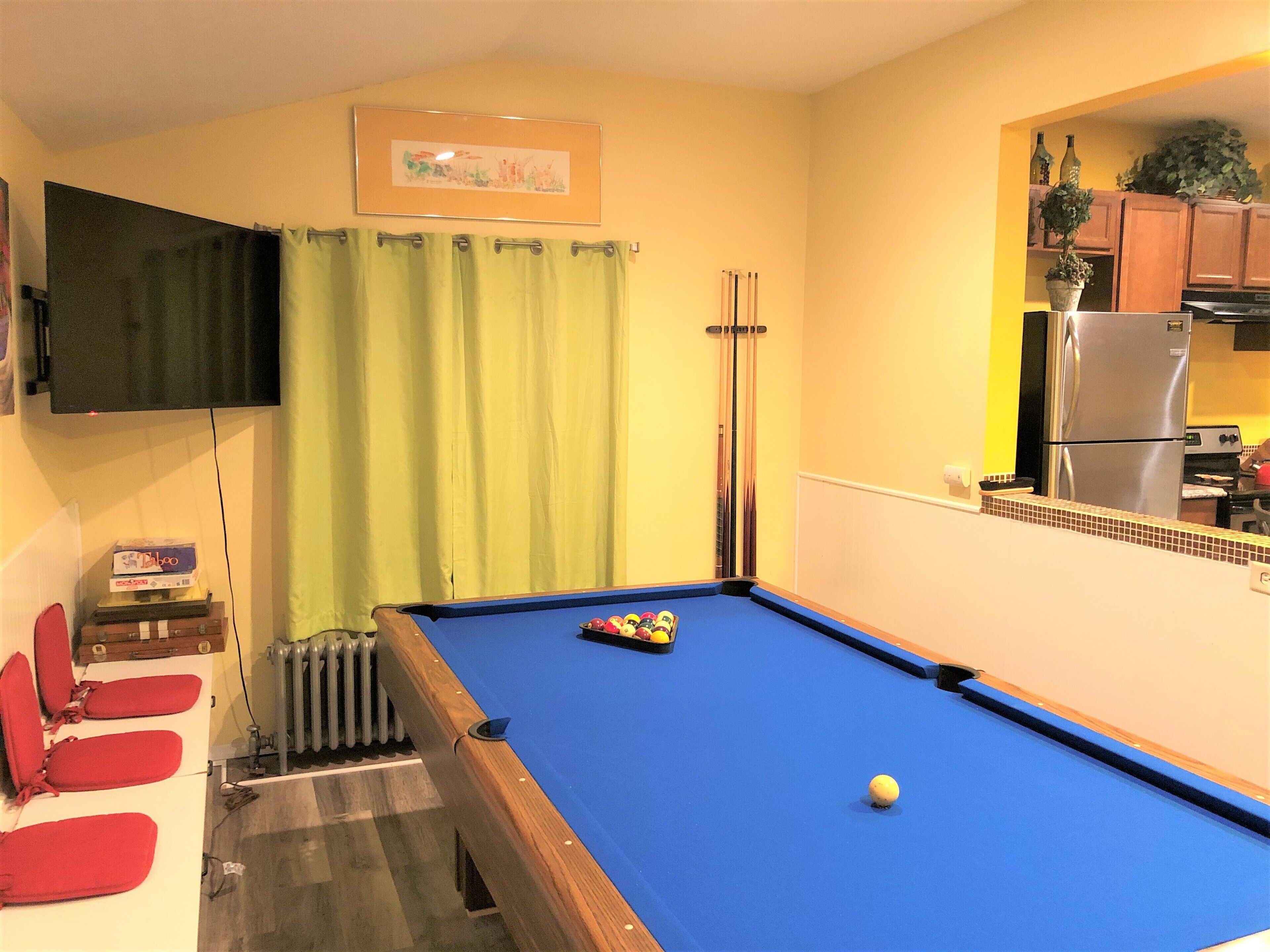 A pool table with a blue felt surface, a yellow wall, a green curtain, and a kitchen area with a refrigerator.