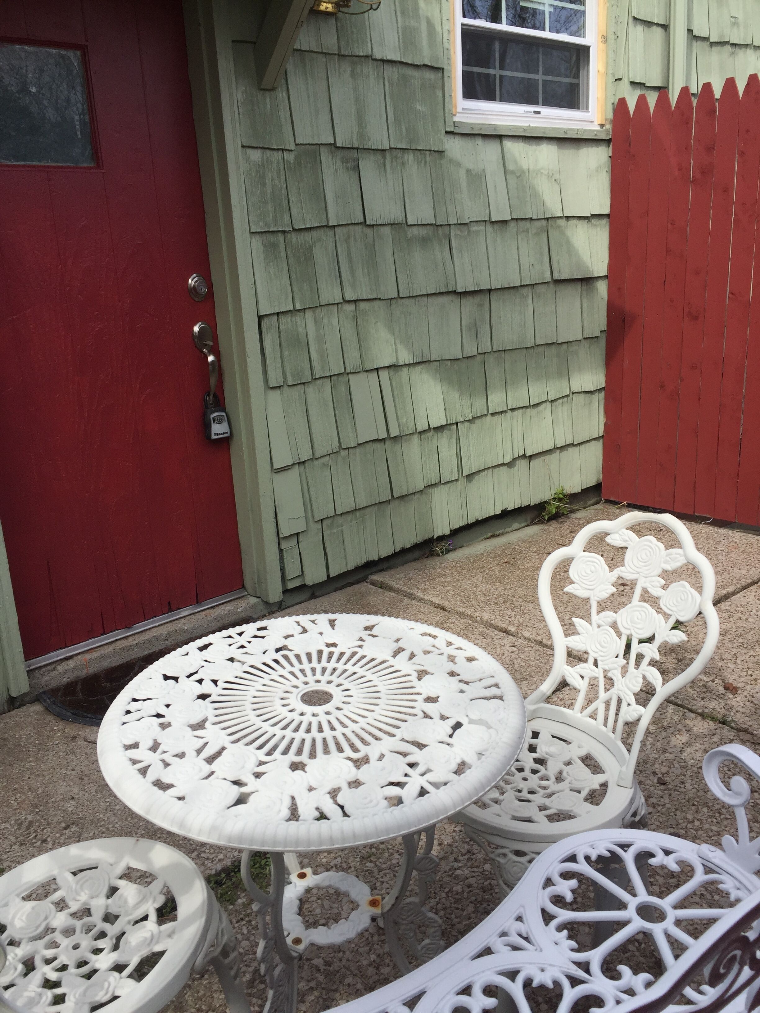 Une petite terrasse avec une table ronde et deux chaises, une porte rouge et un bâtiment aux bardeaux verts.