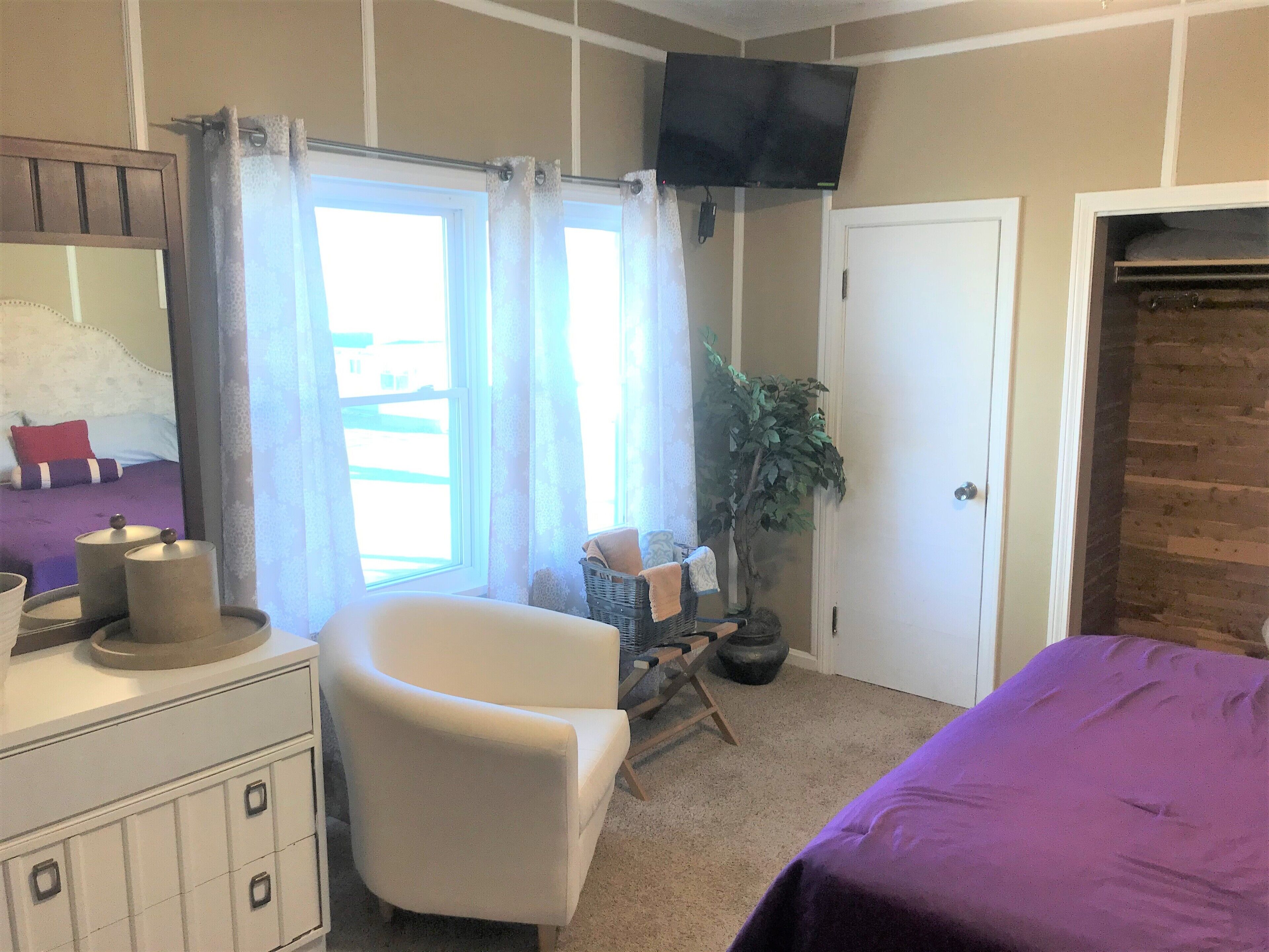 Un dormitorio con cama, una silla, tocador, televisor y ventana con cortinas.