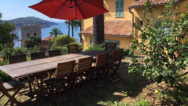 Een terras met een tafel en stoelen, een rode parasol, en uitzicht op zee en een heuvel op de achtergrond.