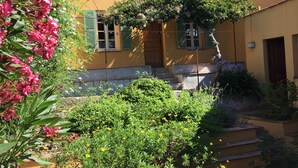 Een gebouw met twee verdiepingen, een gele gevel, groene luiken en een tuin met verschillende planten en bloemen.