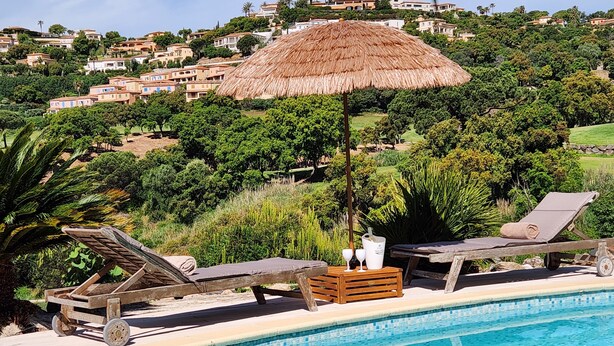 Zona de piscina con sillones de descanso, una sombrilla de paja y vistas a casas en una colina.
