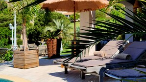 Un área junto a la piscina con sillas de descanso, una sombrilla de paja y plantas tropicales.