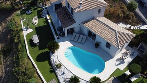 Una casa de dos pisos con piscina, un patio con sillones de descanso y un césped bien cuidado.