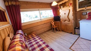 A cozy cabin room with a wooden interior, a plaid blanket on a sofa, a desk with a computer, and a refrigerator.