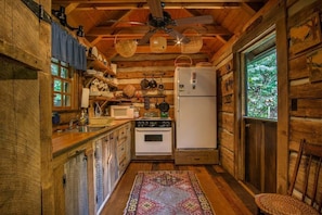 Cocina rústica de cabaña con paredes de madera, ventilador de techo, refrigerador, horno y ventana.