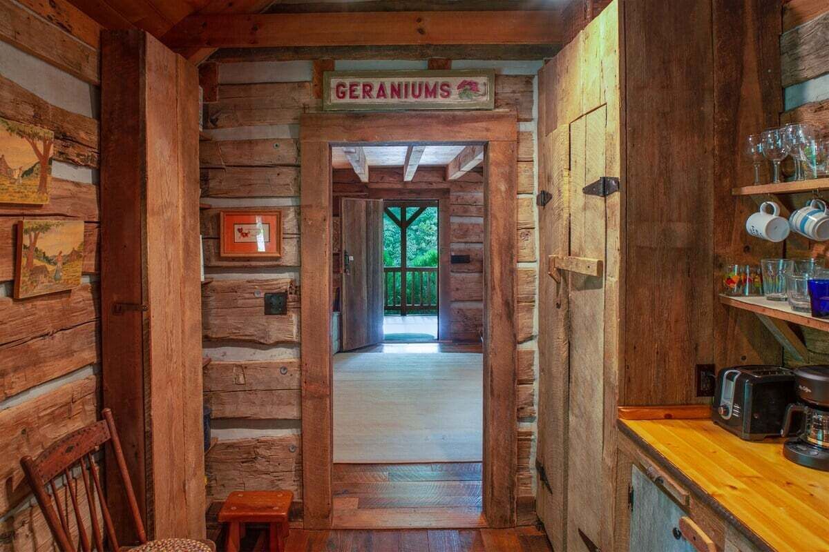 A rustic cabin interior with a wooden door labeled 'GERANIUMS,' a wooden bench, a small table, and a shelf with glasses and a coffee maker.