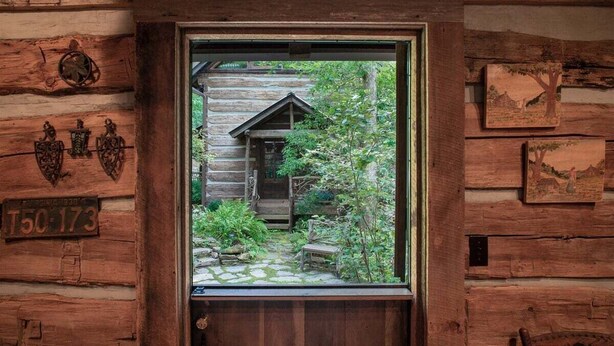 A wooden cabin with a window revealing a stone pathway and a small building in the background. - Aunt Babbies Cabin in Pisgah Forest (Pisgah Forest)