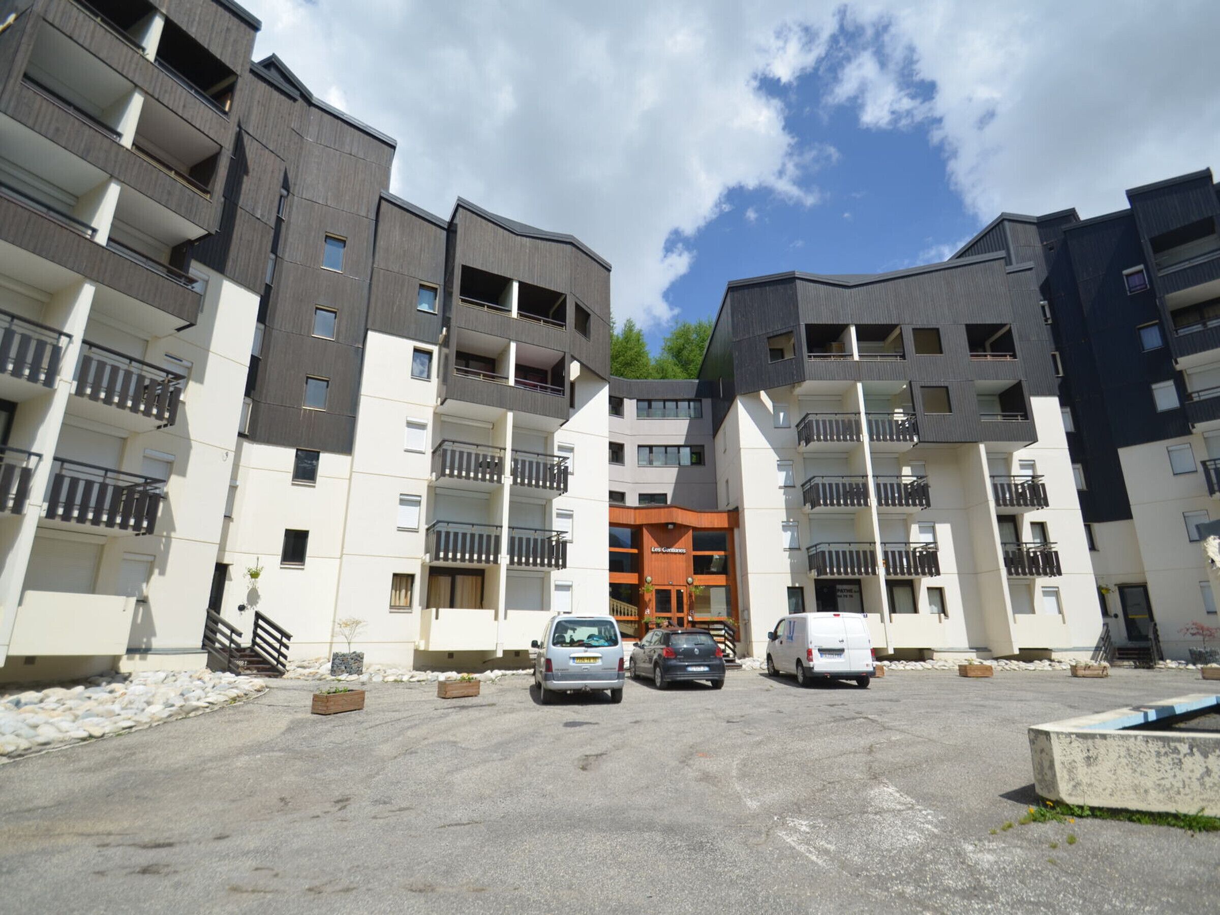 A multi-story residential building with balconies, a central entrance, and a parking area.
