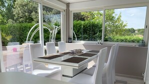 A modern outdoor dining area with a glass table and white chairs, surrounded by large windows offering a view of lush greenery. - Magnificent charming villa with private pool in Sorgues 10 minutes from Avignon  (Sorgues)