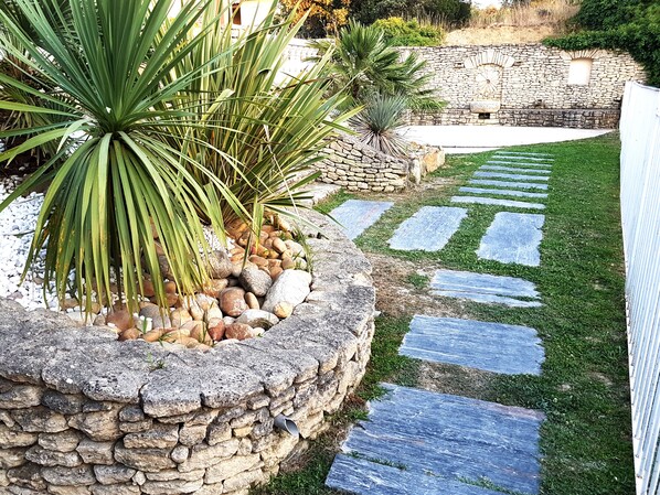 Un sendero de piedra que atraviesa un jardín con palmeras y un muro de piedra al fondo.