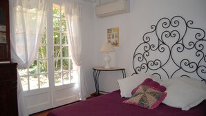 A room with a bed, a window with curtains, a wall-mounted air conditioner, and a small table with a lamp.