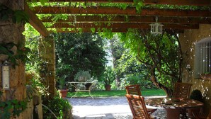 An outdoor patio with wooden furniture, a stone floor, and a pergola covered in greenery.