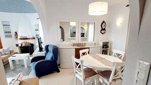 A modern living room with a blue armchair, a white dining table with chairs, and a kitchen area with a white countertop and a hanging light fixture. - Villa Robert. Smart detached Villa with communal pool and fully fenced garden (Calpe)