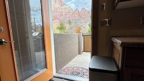 A hotel room entrance with a door leading to a balcony, a trash bin, and a kitchen area with cabinets.