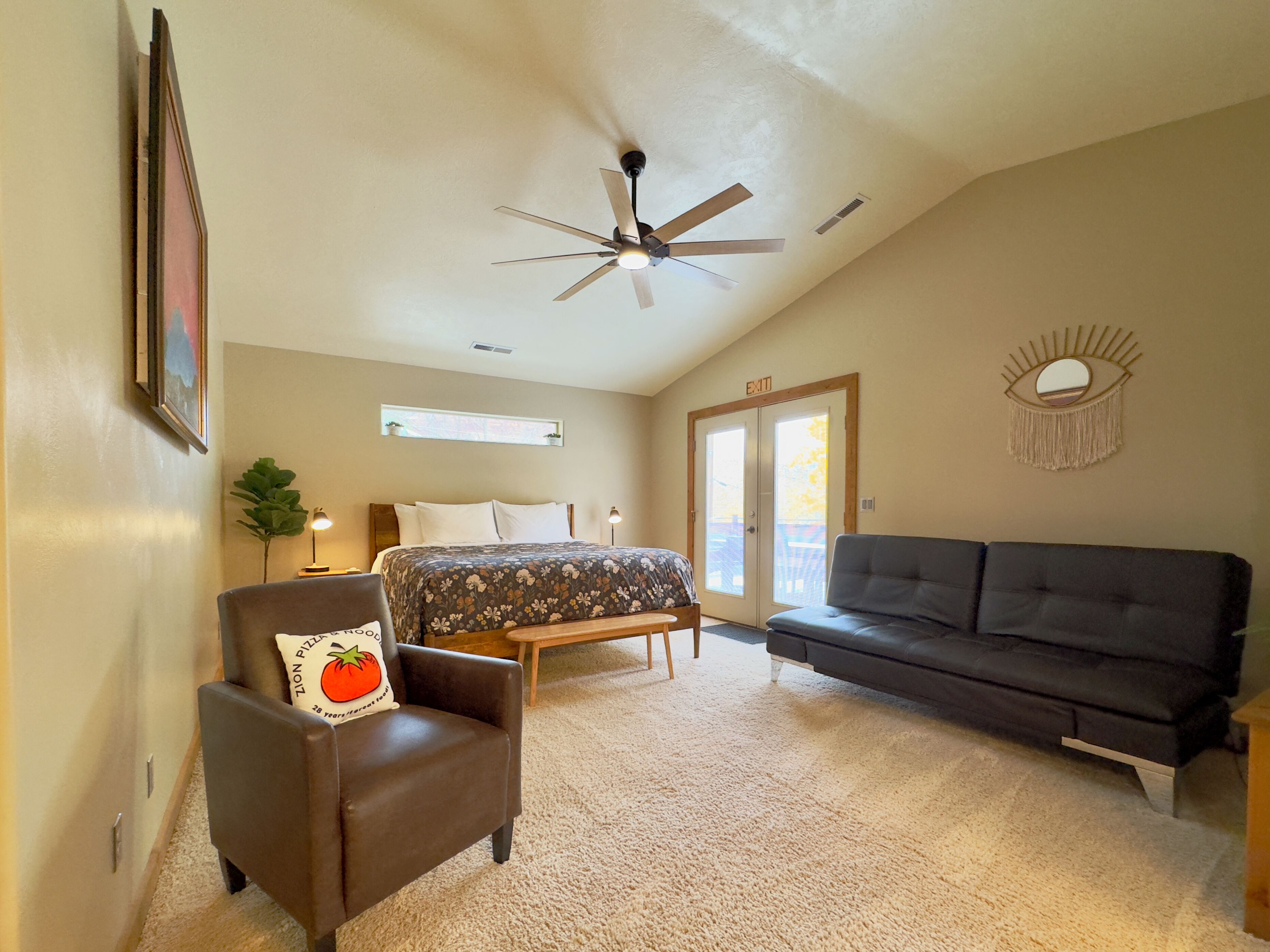 A spacious bedroom with a bed, a dark sofa, a chair, a ceiling fan, and a large window.