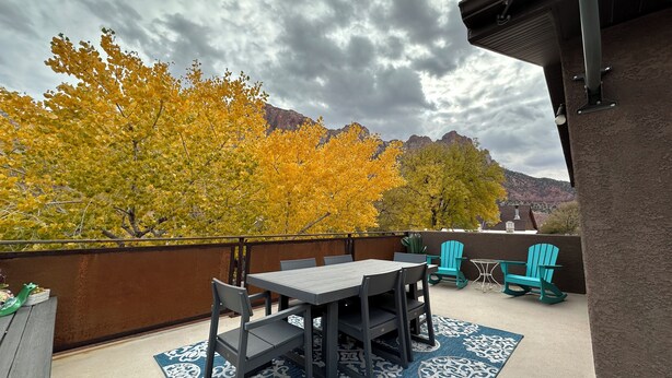 An outdoor patio with a table and chairs, a blue patterned rug, and autumn-colored trees in the background.