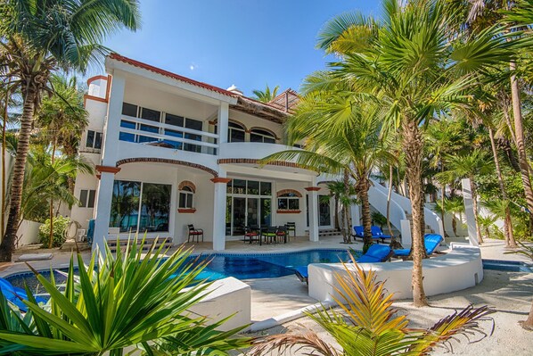 A two-story house with a swimming pool, surrounded by palm trees and a clear blue sky.