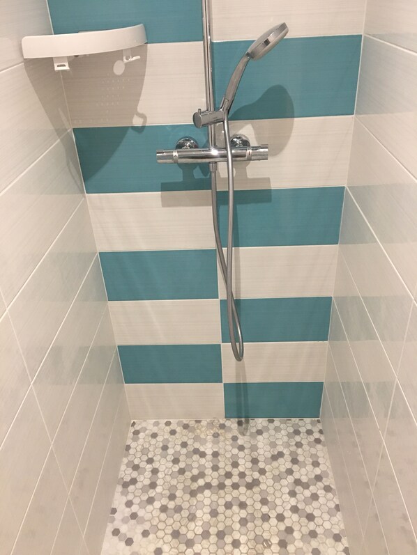 Une douche dotée d’un flexible chromé et de commandes murales, dont les murs sont recouverts de carreaux bleus et blancs formant des rayures, et dont le sol est composé de carreaux hexagonaux.