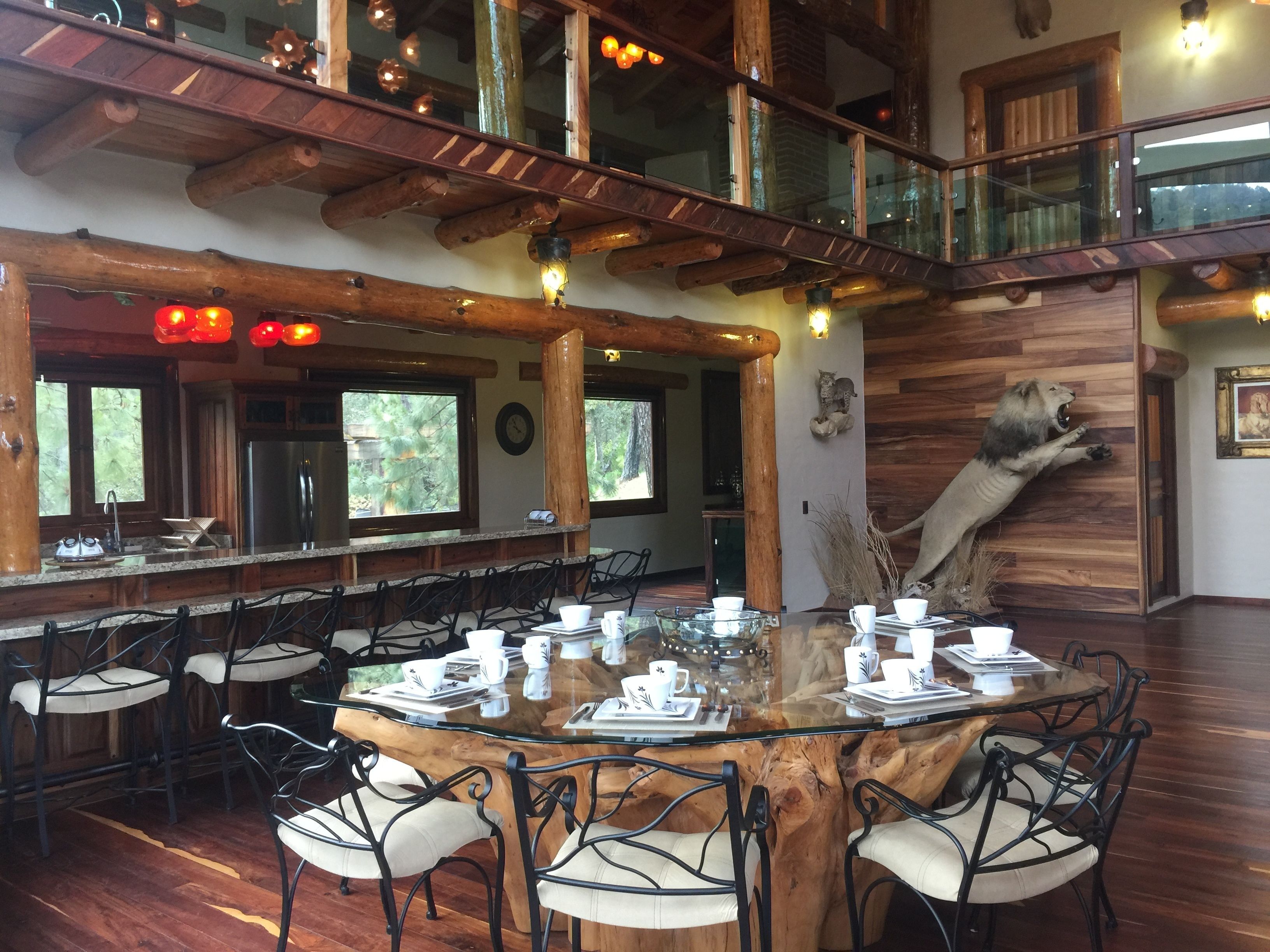 A spacious dining area with a large wooden table surrounded by chairs, a high ceiling with exposed beams, and large windows offering views of the outdoors.