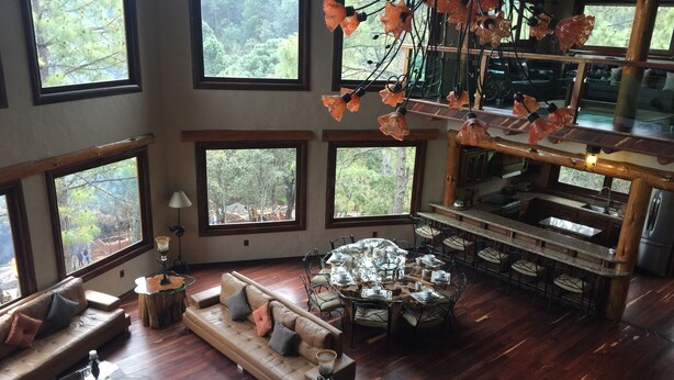 Amplia sala de estar con una gran mesa redonda de vidrio dispuesta para comer, múltiples opciones de asientos y ventanas grandes que ofrecen vistas del bosque circundante.
