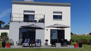 Un bâtiment moderne à deux étages doté d’un balcon, d’un espace extérieur aménagé avec des chaises et des parasols, et d’un barbecue.
