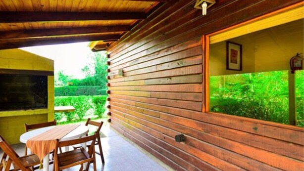 A wooden cabin with a dining area featuring a table and chairs, a window with a view of greenery, and a hanging light fixture.