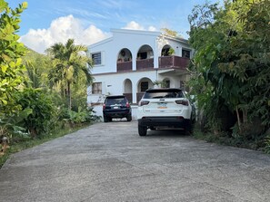 A white building with arched windows and balconies, two parked SUVs, and lush greenery surrounding the property.
