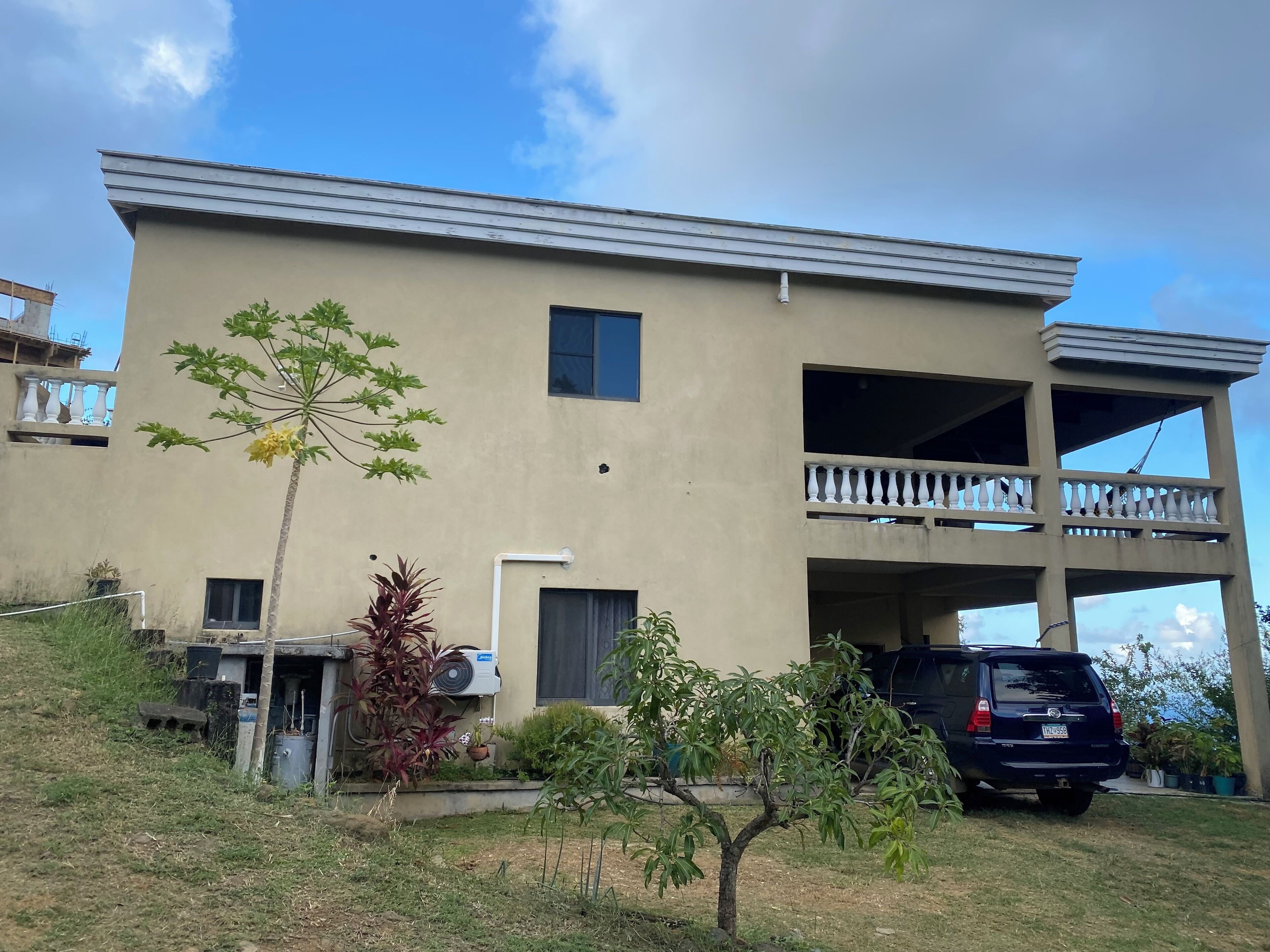 Een gebouw met twee verdiepingen, een balkon, een geparkeerde auto en beplanting in de voortuin.