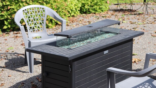 A black outdoor table with a glass top, surrounded by white plastic chairs, set on a gravel surface with green foliage in the background.