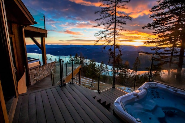 Une terrasse en bois dotée d’un jacuzzi et d’une balustrade en verre, offrant une vue sur un paysage montagneux au coucher du soleil.