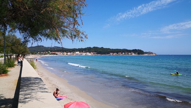 A beach with a concrete walkway, people relaxing under umbrellas, and a kayaker in the water.