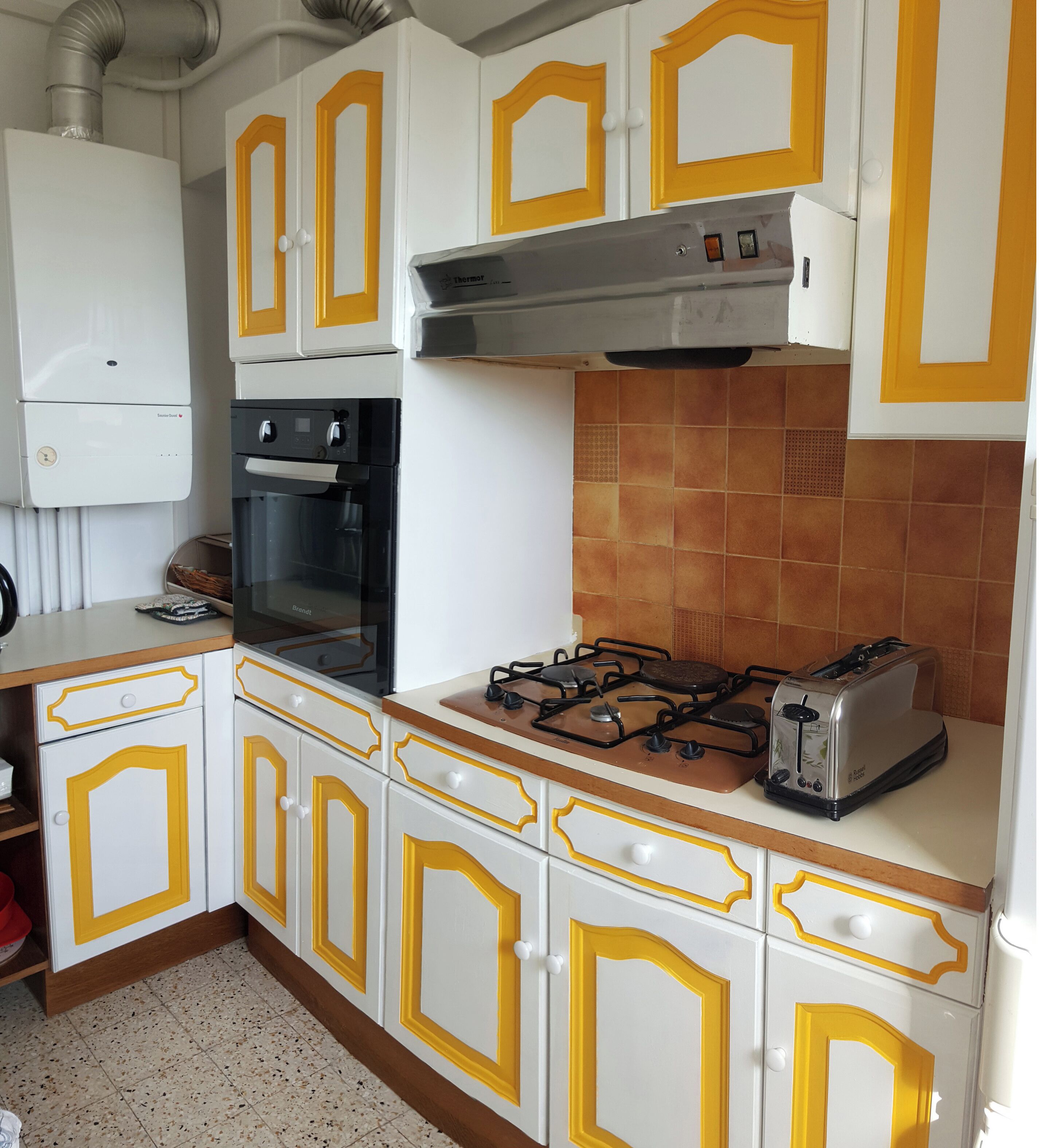 Uma cozinha com armários brancos e detalhes amarelos, fogão a gás, torradeira e forno embutido.