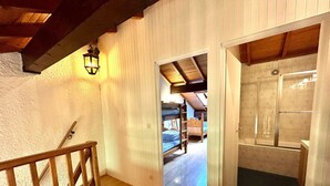 Un escalier en bois avec une rampe, un mur blanc à la finition texturée et une porte donnant sur une pièce où se trouve un lit superposé et une échelle.