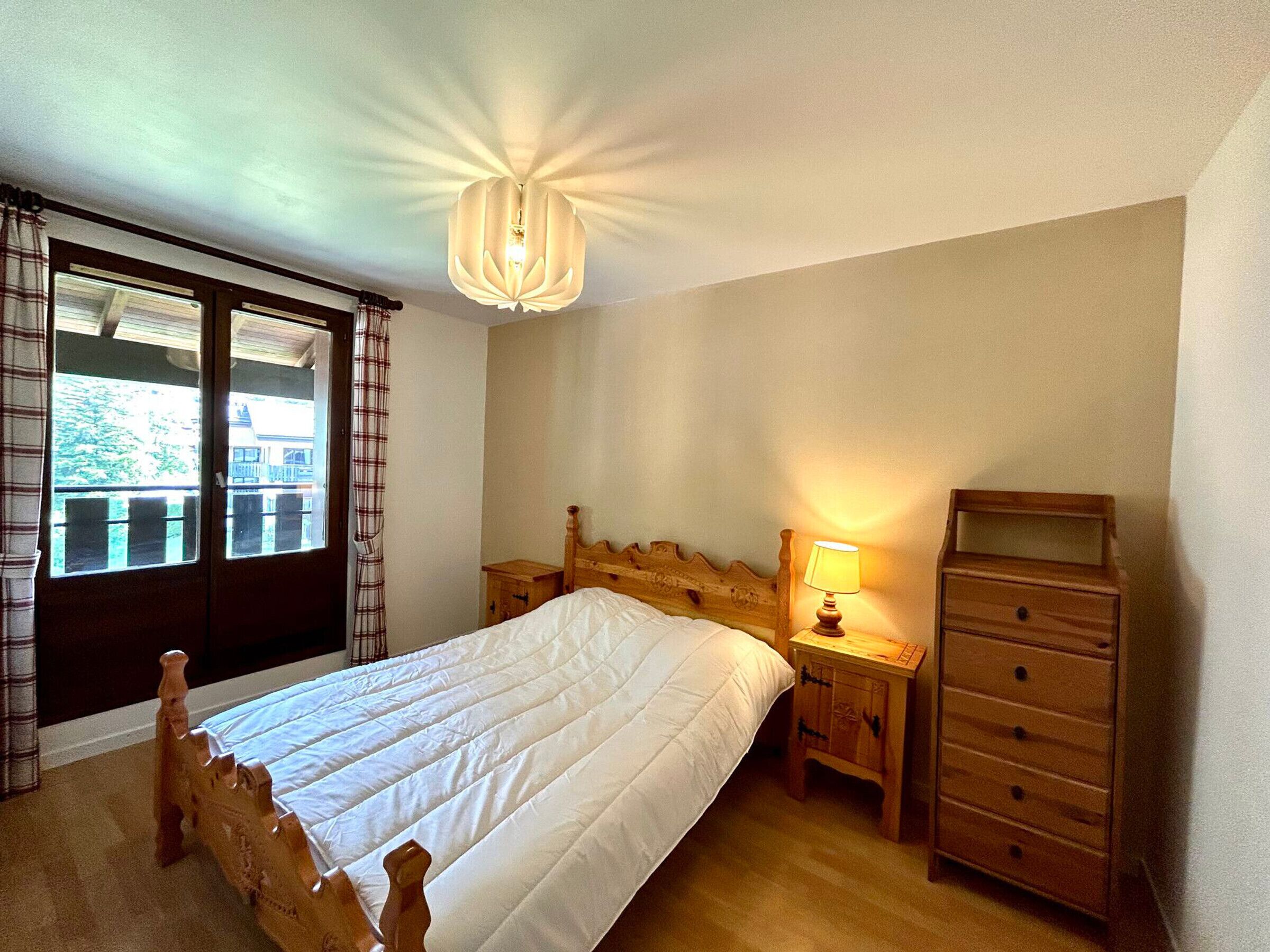 Un dormitori amb un llit de fusta, una tauleta de nit amb una làmpada, un prestatge i una finestra amb cortines.