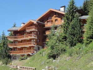 A multi-story wooden chalet with balconies, set in a mountainous area with dense greenery.