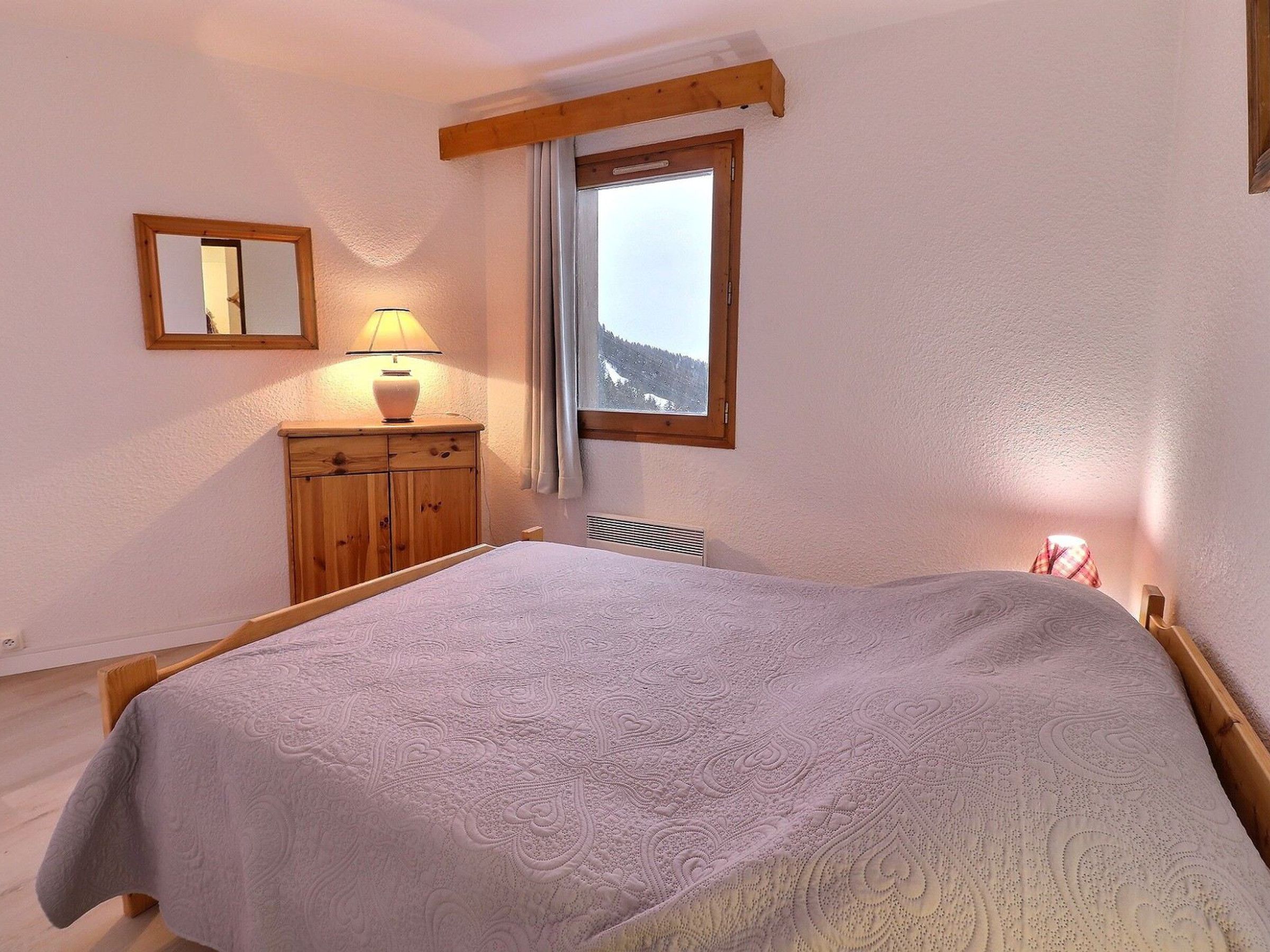 A bedroom with a bed, a wooden dresser, a lamp, a window with a view of mountains, and a mirror on the wall.