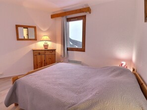 A bedroom with a bed, a wooden dresser, a lamp, a window with a view of mountains, and a mirror on the wall.