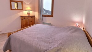 Ein Schlafzimmer mit einem Bett, einer Holzkommode, einer Lampe, einem Fenster mit Bergblick und einem Spiegel an der Wand.