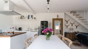 Une cuisine moderne avec des meubles blancs, un four encastré et une machine à café. On y trouve un coin repas avec une table et des chaises blanches, ainsi qu'un vase rempli de fleurs.