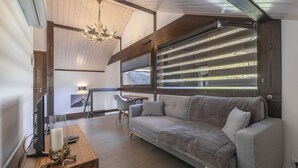 A cozy living room with a sofa, a wooden coffee table, a chandelier, and a large window with blinds. - Le Bois Joly, on track, outdoor spa, sauna (Saint-Gervais-Les-Bains)