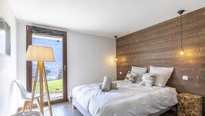 A bedroom with a bed, a standing lamp, a chair, and a view of a cityscape through a window. - Le Bois Joly, on track, outdoor spa, sauna (Saint-Gervais-Les-Bains)