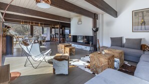 A spacious living area with a large sofa, wooden stools, and a dining table with chairs. There is a fireplace and a large window providing natural light. - Le Bois Joly, on track, outdoor spa, sauna (Saint-Gervais-Les-Bains)