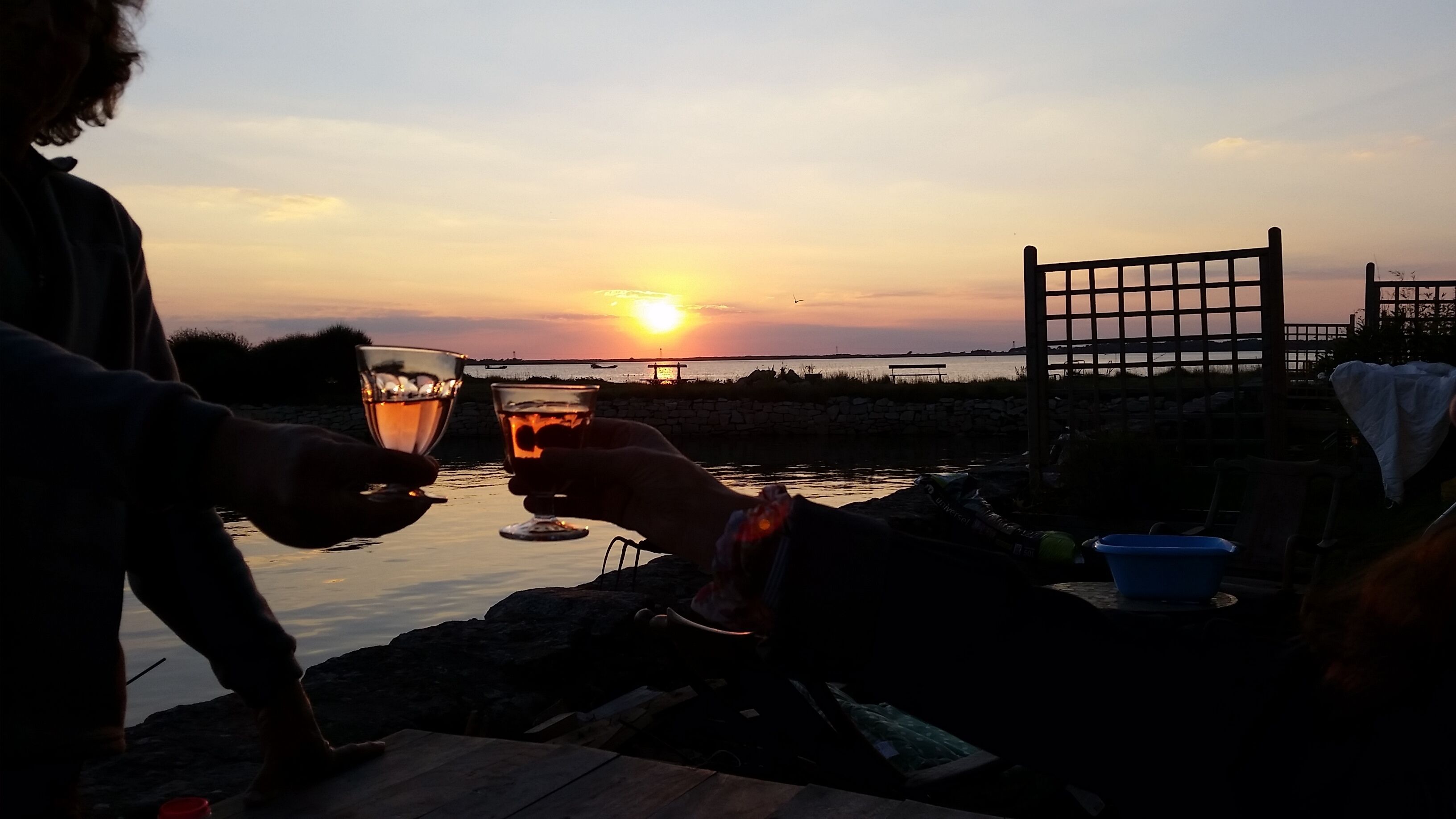 Ein Sonnenuntergang mit zwei Personen, die mit Getränken anstoßen, einem Stuhl und einem Tisch am Wasser.