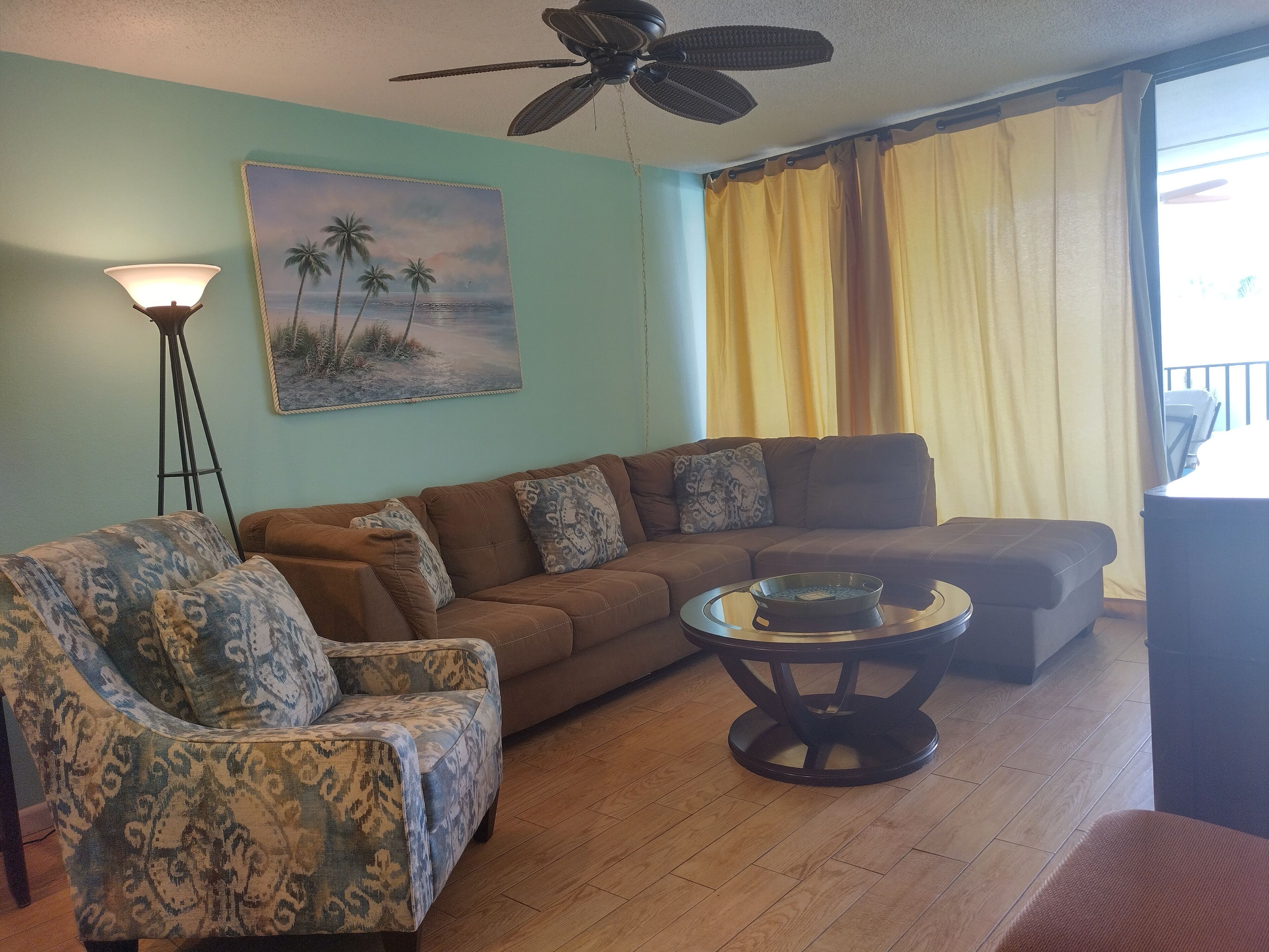 A living room with a brown sofa, patterned armchair, and a coffee table. A painting of palm trees is on the wall, and a ceiling fan is visible.