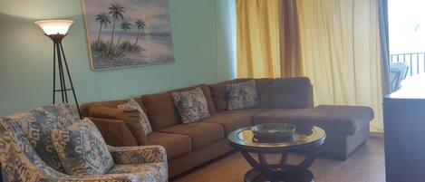 A living room with a brown sofa, patterned armchair, and a coffee table. A painting of palm trees is on the wall, and a ceiling fan is visible.