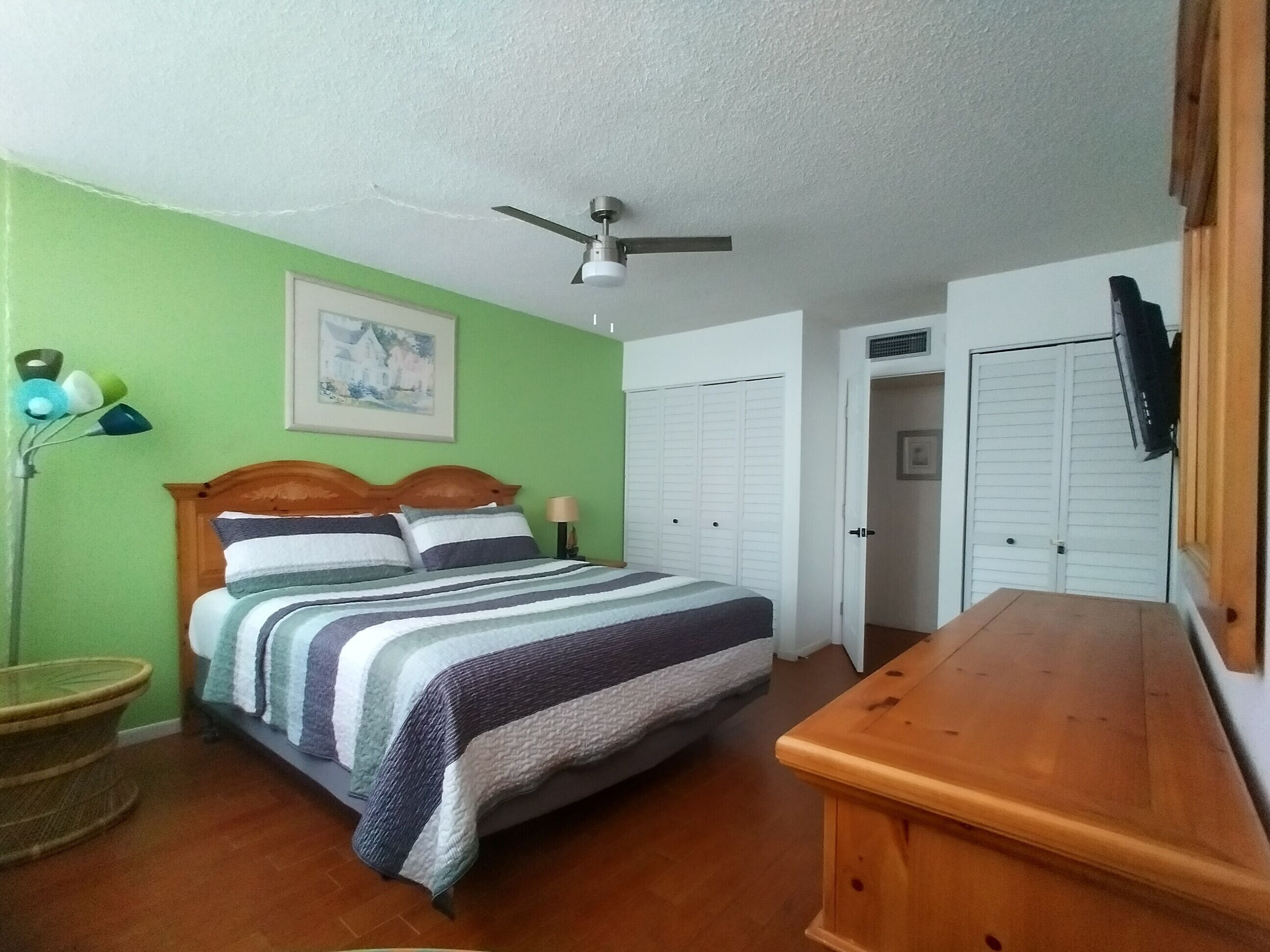 A bedroom with a bed, a ceiling fan, a wooden nightstand, a lamp, and a painting on the wall.