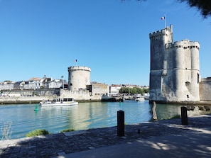 A historic stone castle with a round tower and a tall square tower, situated by a calm river with boats.